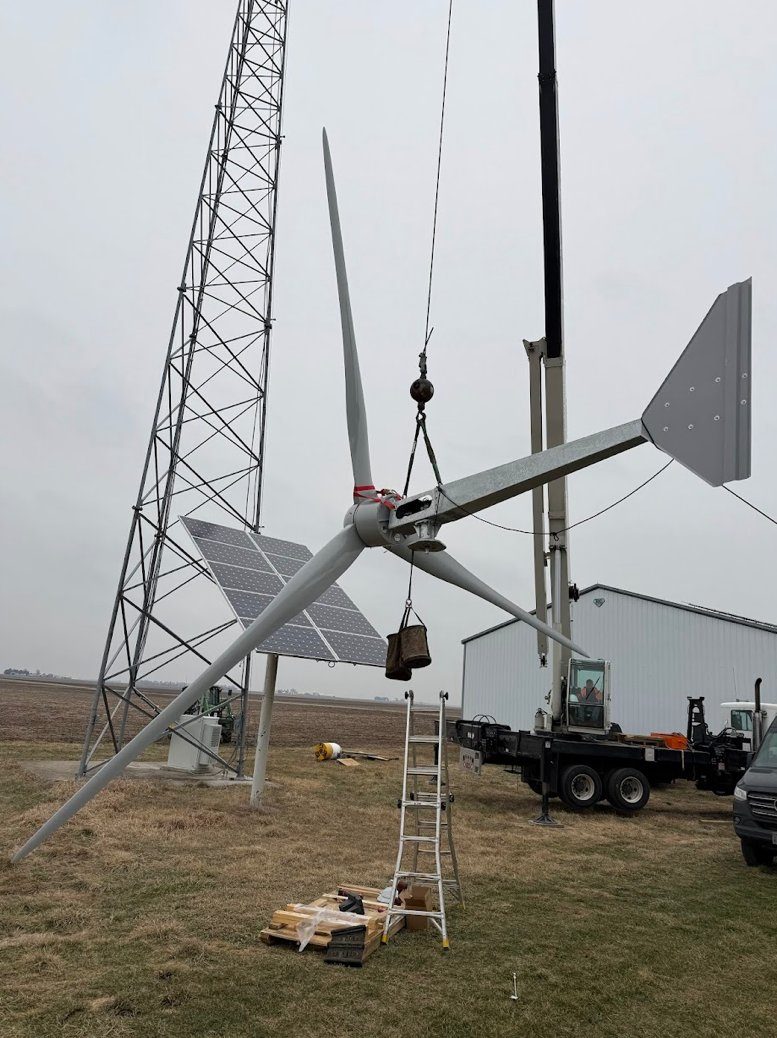 A distributed wind turbine being serviced with a crane on a rural site, with solar panels and a met tower nearby