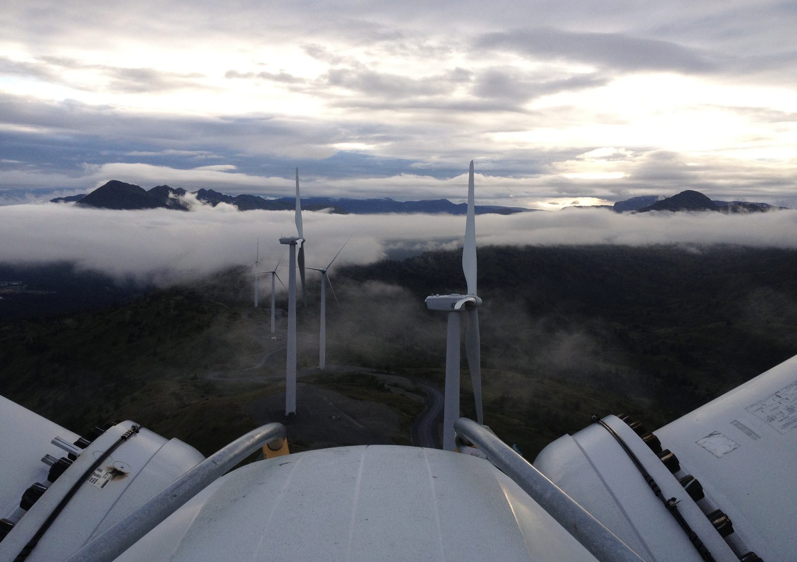 View from the top of a utility-scale wind turbine nacelle, overlooking a mountain wind farm above the clouds